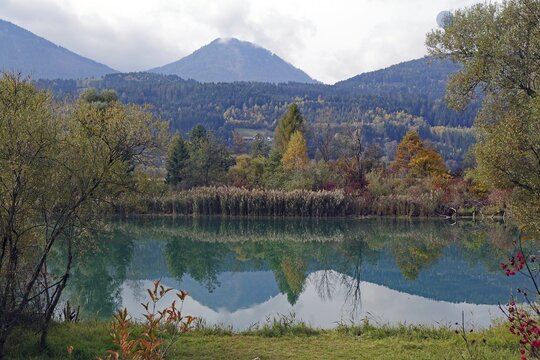 Quiet lake with turquoise-green water, autumnal shore and mountain scenery under cloudy sky, Drau, Drauradweg, Carinthia, near Villach, Austria