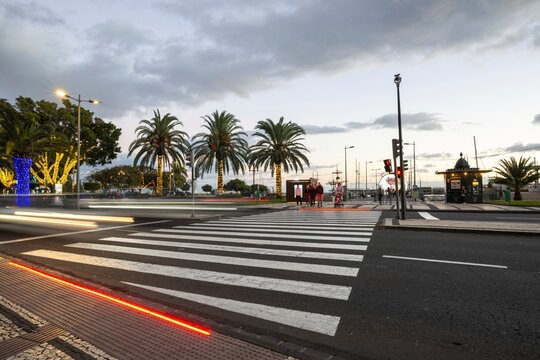 Evening scene at an illuminated pedestrian crossing with integrated LED floor marking, flanked by palm-lined streets and a brightly lit waterfront with Ferris wheel and pavilion. Funchal, Madeira, Portugal
