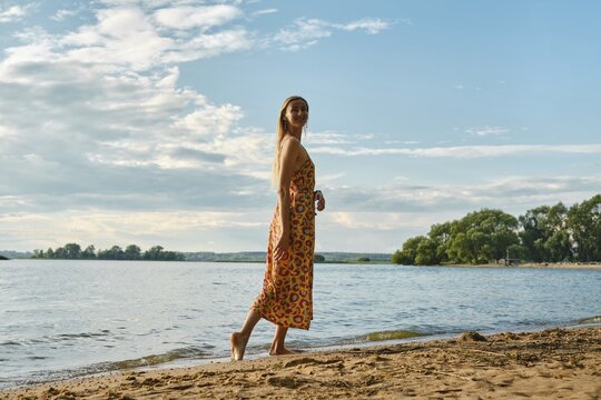 A woman is strolling barefoot along a sandy riverbank, wearing a colourful floral sundress. The blue sky and soft clouds create a serene atmosphere as the sun sets over the water