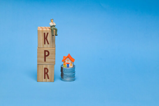 Tiny figure sits atop stacked wooden blocks spelling KPR" beside a miniature house on stacked coins on a bright blue background