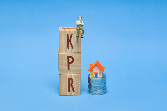 Tiny figure sits atop stacked wooden blocks spelling KPR" beside a miniature house on stacked coins on a bright blue background