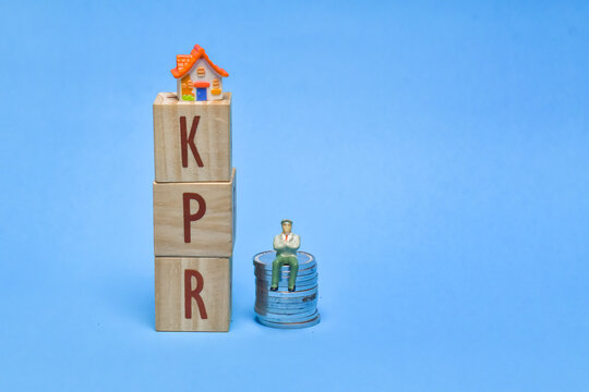 Tiny figure sits atop stacked wooden blocks spelling KPR" beside a miniature house on stacked coins on a bright blue background