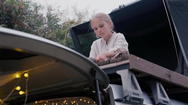 Young girl reading a book sitting on top of a car tent during daylight