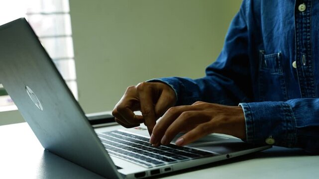 Person Typing on Laptop at Desk, Close-up of a person in a blue denim shirt typing on a laptop at a desk near a window, showing remote work and online communication.