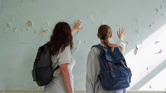 Two people wearing backpacks stand facing weathered wall, peeling paint visible, reaching up touching deteriorating surface while bright sunlight streams across creating strong shadows throughout.