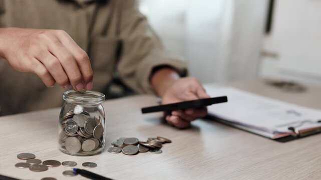 A person puts coins into a glass jar and holds a smartphone beside paperwork on a wooden desk to manage savings, budgeting, financial planning, and micro investment indoors at home.