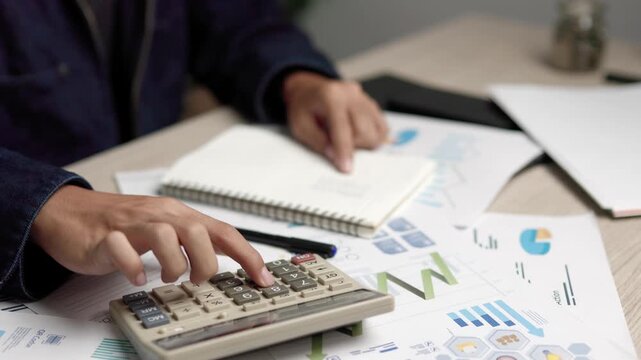 A person calculates numbers with a calculator and writes notes into a notebook beside financial charts and business reports on a wooden desk to analyze profit and budget indoors.