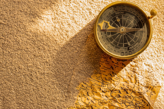 Vintage brass compass and old map partly buried in sand in sunlight background backdrop
