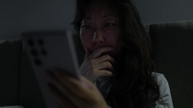 Frightened woman reading shocking news on smartphone at night. Asian woman lying on bed at home at night, holding her smartphone and reacting with shock and fear to something she reads on the screen.