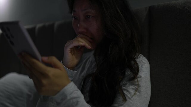 Frightened woman reading shocking news on smartphone at night. Asian woman lying on bed at home at night, holding her smartphone and reacting with shock and fear to something she reads on the screen.