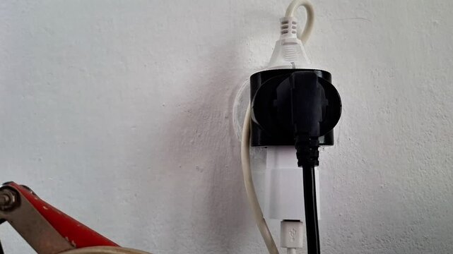 Black electrical plug adapter connected to a white wall socket, with power cables hanging against a plain wall, illustrating household electricity, electrical connections, and everyday power supply