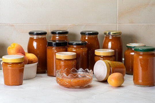 A collection of jars filled with homemade apricot jam is displayed on a kitchen countertop. The jars vary in size and shape, with some featuring colorful lids.