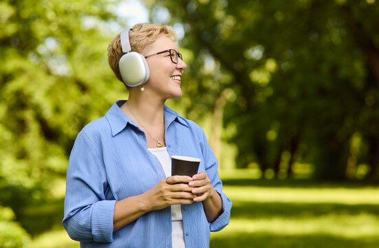 Senior woman with headphones and coffee enjoying music. Smiling in a green park, she relaxes outdoors with a warm drink and relaxation vibe. Positive wellbeing and leisure lifestyle concept.