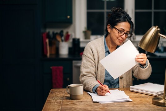 Middle aged woman with glasses sitting at wooden table in kitchen at night reading paper document and writing with pen under desk lamp
