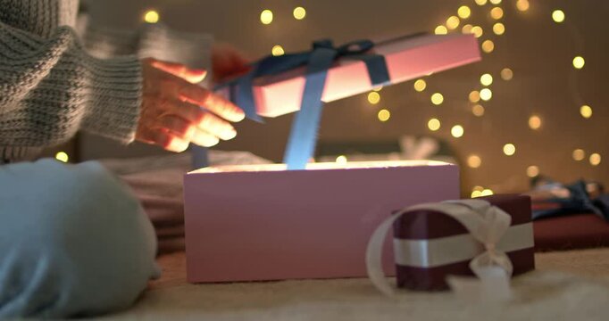 Woman hands opening gift box with glowing light inside looking into it on bokeh garland background close up video
