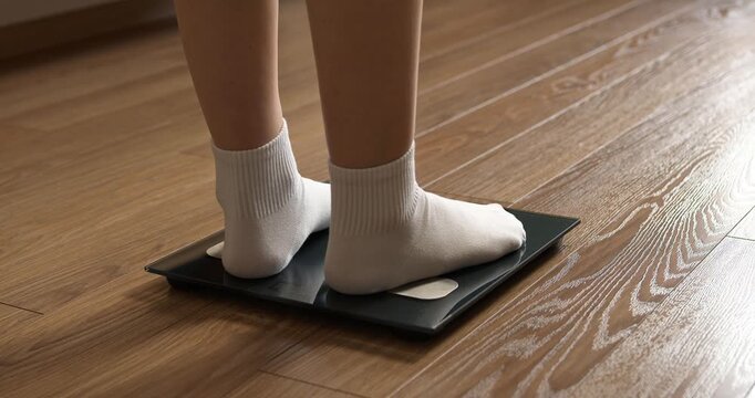 Woman feet in white socks stepping on digital electronic scales to check weight on wooden floor.