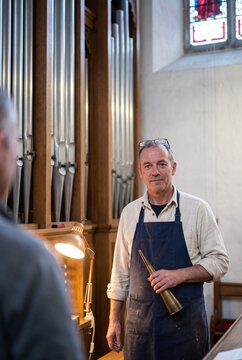 Skilled craftsman and organ builder holding a brass tuning cone while standing next to a large traditional church pipe organ during maintenance work