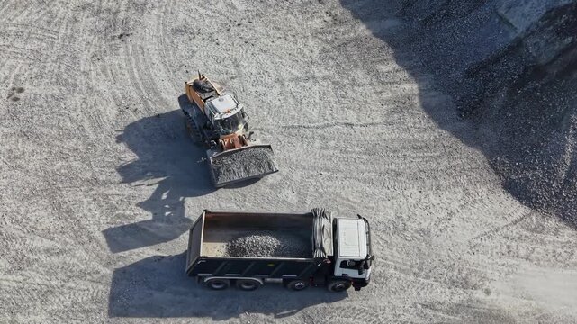 A loader moves gravel across a quarry work area while a dump truck waits nearby during daytime operations.