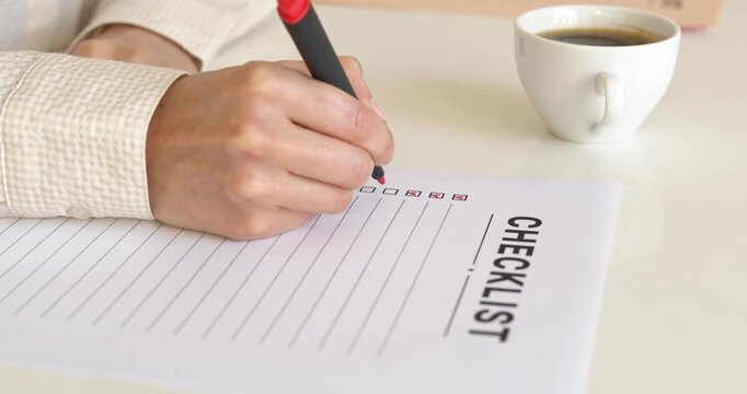 Woman hand checking off boxes with red marker on paper checklist next to coffee cup.