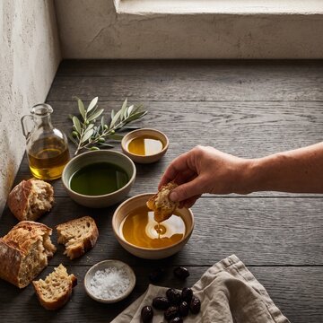 Person dipping crusty artisan bread into a ceramic bowl of golden olive oil on a rustic dark wooden table with olives and salt
