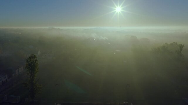 Aerial video across a shallow layer of ground fog and smoke drifting over a small farming community in serbian town of Temerin, Vojvodina. The sun sits low on the horizon at sunrise