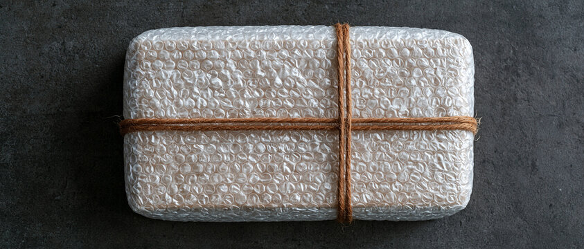 Top view of a securely wrapped package in clear bubble wrap, tied with rustic brown twine on a dark, textured concrete background, emphasizing safe delivery and careful handling of fragile