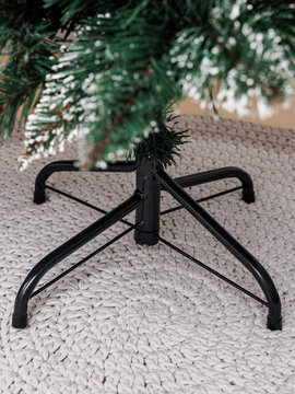 Black metal stand supports artificial pine tree on woven rug in room. This detailed shot captures technical preparation for New Year celebration.