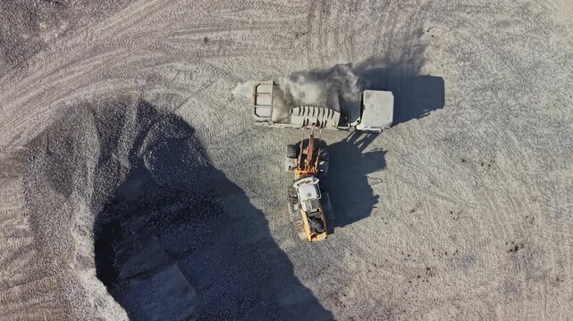 At a gravel site, a loader works beside a dump truck, moving material into its bed during daytime as seen from above.