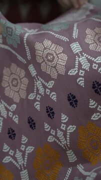 Cinematic close-up footage of a hand holding traditional Indonesian woven songket fabric with colorful floral motifs on purple textile.