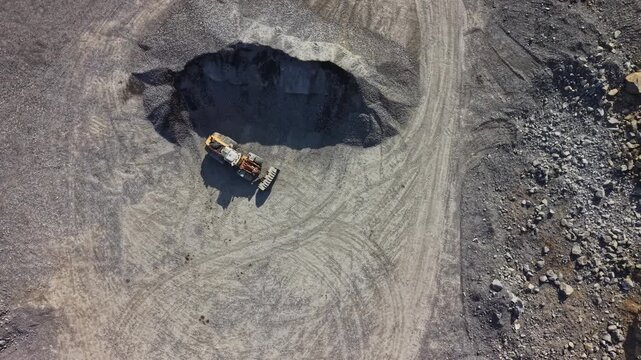 A worker drives a loader beside a large pit at an open pit mine, with tire tracks and rock visible across the work area in daylight.