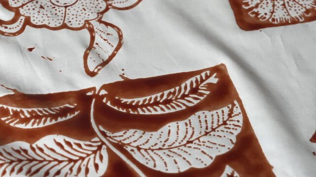 Macro cinematic detail footage of traditional Indonesian batik with brown wax resist forming intricate leaf and floral patterns on white cotton fabric, close-up handmade textile texture.