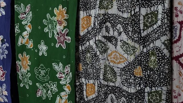 Cinematic close-up footage of colorful finished Indonesian batik fabrics hanging side by side, displaying traditional floral and abstract patterns in green, black, red and blue.