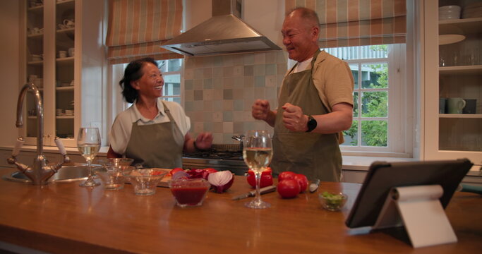 In kitchen, Senior Asian couple dancing surrounded by cooking ingredients and tablet, at home