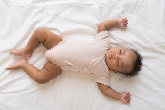 African American newborn baby sleeping soundly on white bed sheets with hands raised up, top view portrait of cute innocent infant napping comfortably in cozy bedroom