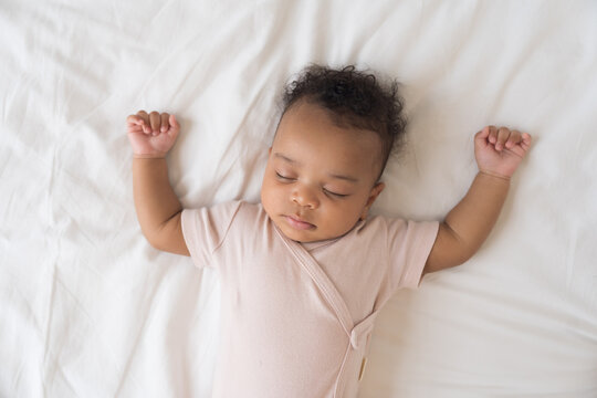 African American newborn baby sleeping soundly on white bed sheets with hands raised up, top view portrait of cute innocent infant napping comfortably in cozy bedroom