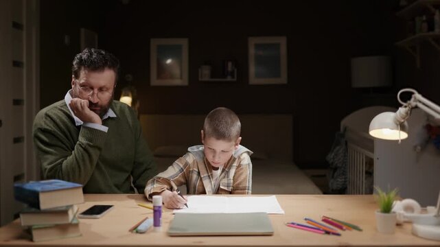 Father watches son doing homework at home in evening. Dad supervises child completing school homework. Parenting supervision education concept