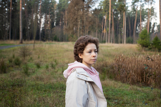 woman spends leisure time at the edge of a pine forest, standing in a light jacket and pink hoodie with a grassy clearing and a paved walking path in the background