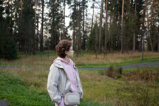 woman with cancer stands beside a paved woodland path during an outdoor walk, looking toward the pine trees with her hands in her pockets and a small bag across her body