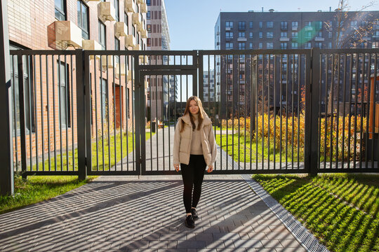 woman in a beige puffer vest walks toward the camera beside a metal courtyard gate, with apartment buildings, sunlit shrubs and long striped shadows across the paved walkway