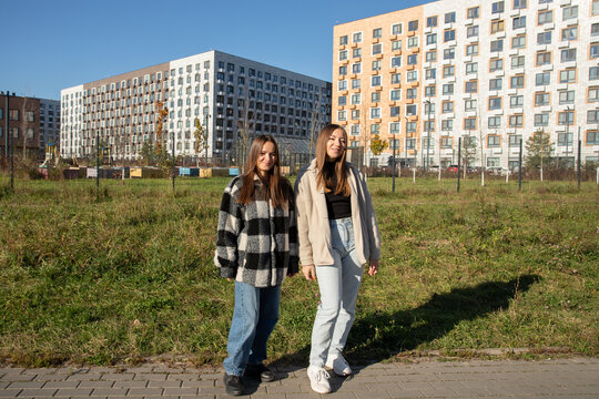 two women pose together on a paved path in beige and plaid jackets with blue jeans, their white and black shoes visible as grass, young trees and apartment buildings fill the background