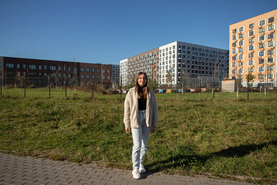 woman stands on a paved path in a residential neighborhood, wearing a beige jacket, pale blue jeans and white sneakers, with apartment buildings and clear blue sky behind her