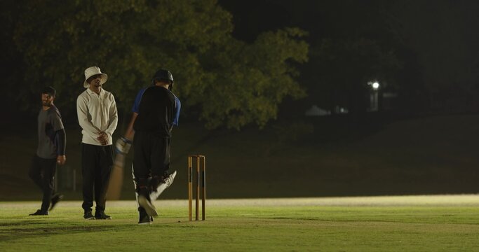 Bowler returning to crease on floodlit field prompts batsman practicing swinging bat, bumping fists