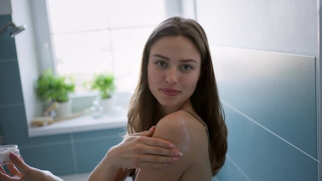 Young single woman doing skincare routine in bathroom on the edge of bathtub.