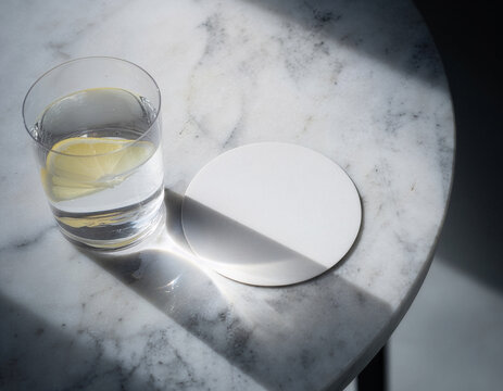 Blank white round coaster mockup beside a glass of lemon water on a sunlit white marble table