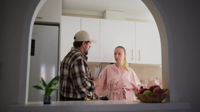 Professional male plumber in cap talking with female homeowner in pink bathrobe in domestic kitchen, holding clipboard to discuss pipe repair service and maintenance estimate