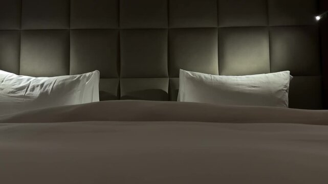 Quiet hotel bed with white pillows and padded headboard illuminated by bedside lights in a dark, cozy bedroom interior