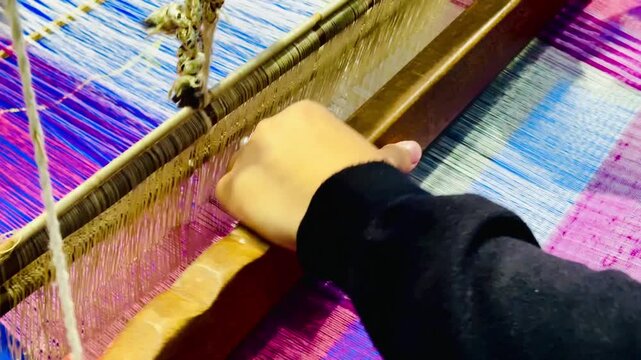 Close-up of hands weaving colorful silk threads on a traditional loom, creating intricate textile patterns with vibrant pink, purple, and blue fibers during the Thai silk weaving process.