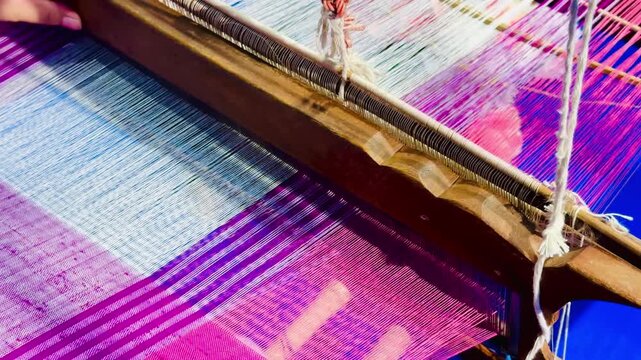 Close-up of hands weaving colorful silk threads on a traditional loom, creating intricate textile patterns with vibrant pink, purple, and blue fibers during the Thai silk weaving process.