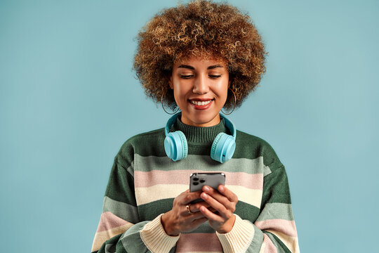 Smiling young woman with curly hair uses a smartphone while wearing blue headphones against a plain light blue studio background.