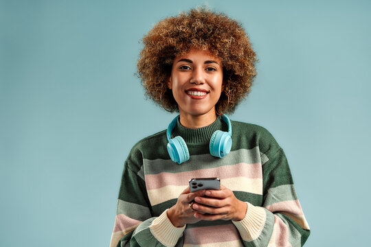 Smiling young woman with curly hair wearing turquoise headphones and holding a smartphone against a soft blue studio background.